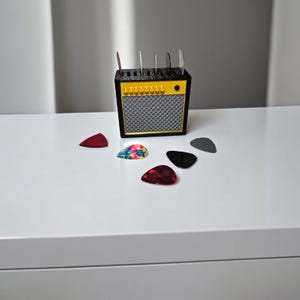 May include: A black and yellow guitar pick holder shaped like a small amplifier. The holder has slots for multiple picks and is surrounded by colourful guitar picks on a white surface.