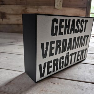 Op de afbeelding: Een zwart ingelijst bord met een witte achtergrond en zwarte tekst die "GEHASST VERDAMMT VERGÖTTERT" luidt.