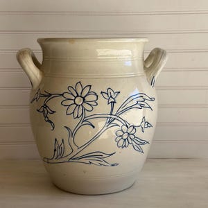 May include: Cream-colored ceramic vase with two handles and a blue floral design. The vase features a rounded body and a slightly flared rim. The blue floral pattern includes flowers, leaves, and stems.