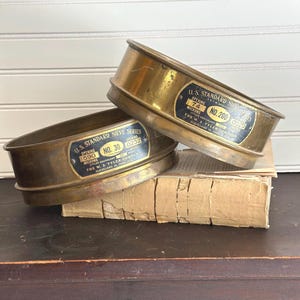 May include: Two vintage brass sieves from the U.S. Standard Sieve Series, with labels indicating micron openings and the manufacturer, The W.S. Tyler Company. One sieve is labeled No. 30, the other No. 200. They rest on a stack of aged paper.