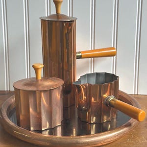 May include: A copper coffee pot, creamer, and sugar bowl set with wooden handles. The set is on a copper tray with a mirrored surface.
