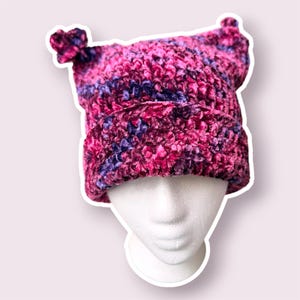 Peut inclure: Un bonnet en velours pelucheux aux teintes vives de magenta et de violet profond. Le bonnet présente un bord replié et deux petites protubérances arrondies en forme d'oreilles sur les coins supérieurs, en fil doux et duveteux multicolore.