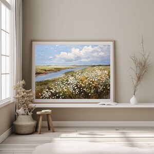 May include: An oil painting depicting a serene landscape with a river, wildflowers, and a blue sky dotted with white clouds. The artwork is framed in a light wood frame and displayed in a neutral-toned room. The painting conveys a sense of peace and natural beauty.