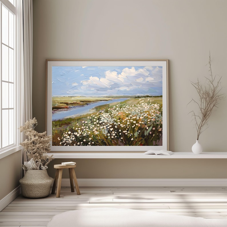 May include: An oil painting depicting a serene landscape with a river, wildflowers, and a blue sky dotted with white clouds. The artwork is framed in a light wood frame and displayed in a neutral-toned room. The painting conveys a sense of peace and natural beauty.