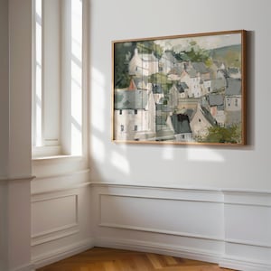 May include: A framed landscape painting depicts a village nestled on a hillside. The artwork features white buildings with gray roofs, surrounded by green foliage. The painting is in a light brown wooden frame, hanging on a white wall.