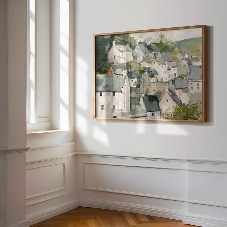 May include: A framed landscape painting depicts a village nestled on a hillside. The artwork features white buildings with gray roofs, surrounded by green foliage. The painting is in a light brown wooden frame, hanging on a white wall.
