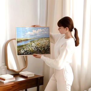 May include: A framed landscape painting depicting a river, field of white flowers, and a blue sky with clouds. The artwork is in a gold frame. The painting is being held up in front of a mirror and a wooden table.