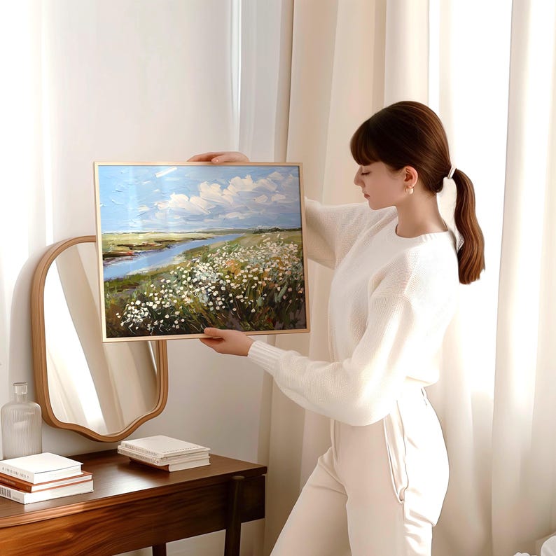 May include: A framed landscape painting depicting a river, field of white flowers, and a blue sky with clouds. The artwork is in a gold frame. The painting is being held up in front of a mirror and a wooden table.