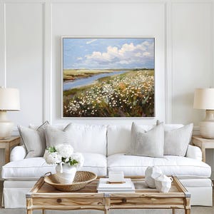 May include: An oil painting depicting a landscape with a river, wildflowers, and a cloudy sky. The artwork is framed and hangs above a white sofa with decorative cushions. A wooden coffee table with decor and lamps complete the scene.