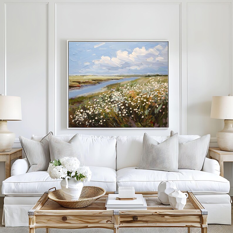 May include: An oil painting depicting a landscape with a river, wildflowers, and a cloudy sky. The artwork is framed and hangs above a white sofa with decorative cushions. A wooden coffee table with decor and lamps complete the scene.