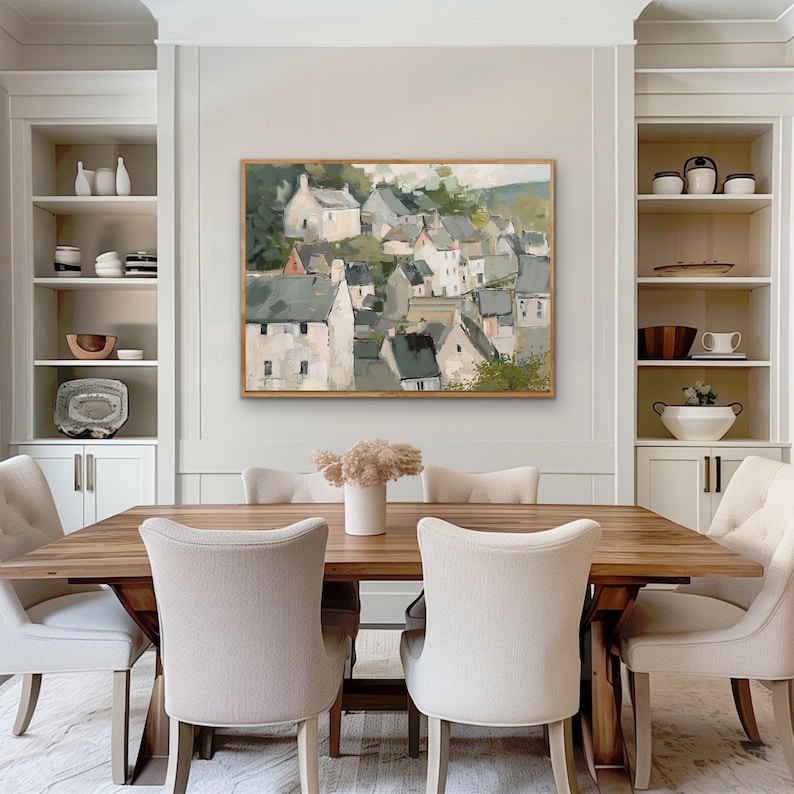 May include: A dining room featuring a large wooden table with six beige upholstered chairs. A framed landscape painting of a village hangs above the table. White built-in shelves on either side display decorative items, creating a cozy and inviting atmosphere.