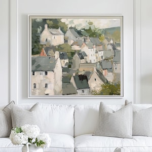 May include: An abstract painting of a village with white buildings and gray rooftops. The artwork is framed in a white square frame. The painting has a neutral color palette with green and beige tones. The artwork is displayed above a white sofa with gray pillows.
