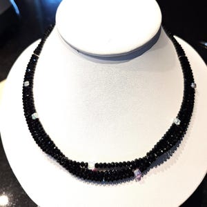 May include: A multi-strand black beaded necklace, presented on a white display bust. The necklace is composed of small, dark beads with clear and pink accent beads. The necklace is designed to be worn around the neck.