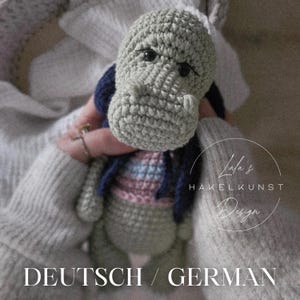 May include: A crocheted hippo doll in shades of gray, blue, and pink. The handmade toy has a detailed snout, small black eyes, and a blue jacket. The image includes the text "DEUTSCH / GERMAN" and the designer's logo.