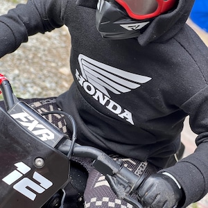 May include: A person wearing a black hooded sweatshirt with a white Honda logo, a black helmet with a red visor, and black gloves. The person is riding a black dirt bike.