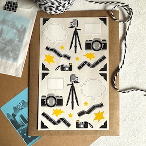 Puede incluir: Una hoja de papel blanco con ilustraciones en blanco y negro de cámaras, rollos de película, estrellas y burbujas de diálogo. Las ilustraciones están dispuestas en un patrón repetitivo.