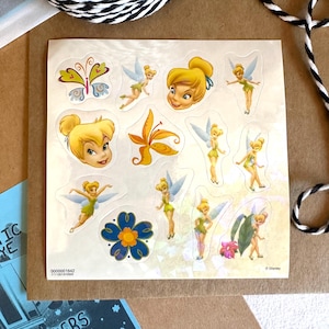 May include: A sheet of stickers featuring Tinker Bell, a fairy with blonde hair and green wings. The stickers are on a white background with a pearlescent finish. The stickers include Tinker Bell's face, Tinker Bell flying, a butterfly, a flower, and a leaf. The text "© Disney" is in the bottom right corner.