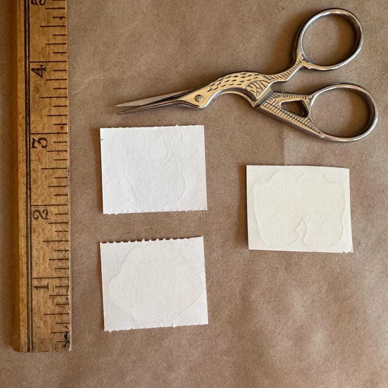 May include: Three white paper squares with a raised design, possibly a flower or leaf, on a brown surface. A pair of silver scissors with a bird-shaped handle is also on the surface.