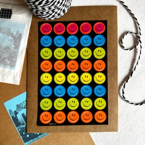 May include: A sheet of colorful smiley face stickers on a black background, arranged in rows and columns. The stickers feature a variety of colors, including pink, blue, green, orange, and yellow. The sheet is on a brown card, with a black and white string and other stationery items.