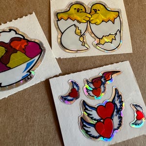 May include: Three sheets of stickers with holographic designs. The first sheet features a colorful ice cream sundae in a bowl. The second sheet features two yellow chicks hatching from eggs. The third sheet features four stickers of red hearts with wings.