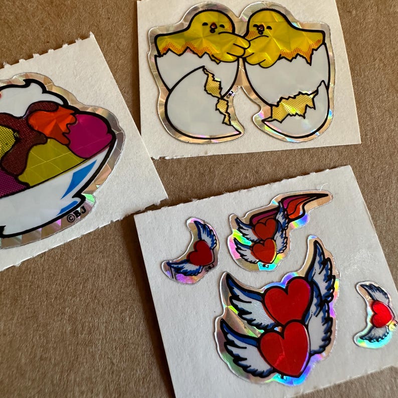 May include: Three sheets of stickers with holographic designs. The first sheet features a colorful ice cream sundae in a bowl. The second sheet features two yellow chicks hatching from eggs. The third sheet features four stickers of red hearts with wings.