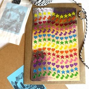 May include: A sheet of gold foil stickers with a rainbow of colors. The stickers feature stars, hearts, and musical notes. The sheet is labeled "Decorate A Finger" and "Sandylon Sticker Designs 1983".