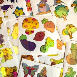 Może przedstawiać: Arkusz kolorowych, holograficznych naklejek z różnymi postaciami i przedmiotami z kreskówek, w tym grzybem, bakłażanem, marchewką, motylem i żabą. Naklejki idealnie nadają się do dodania odrobiny fantazji do dzienników, planerów i innych projektów rękodzielniczych.