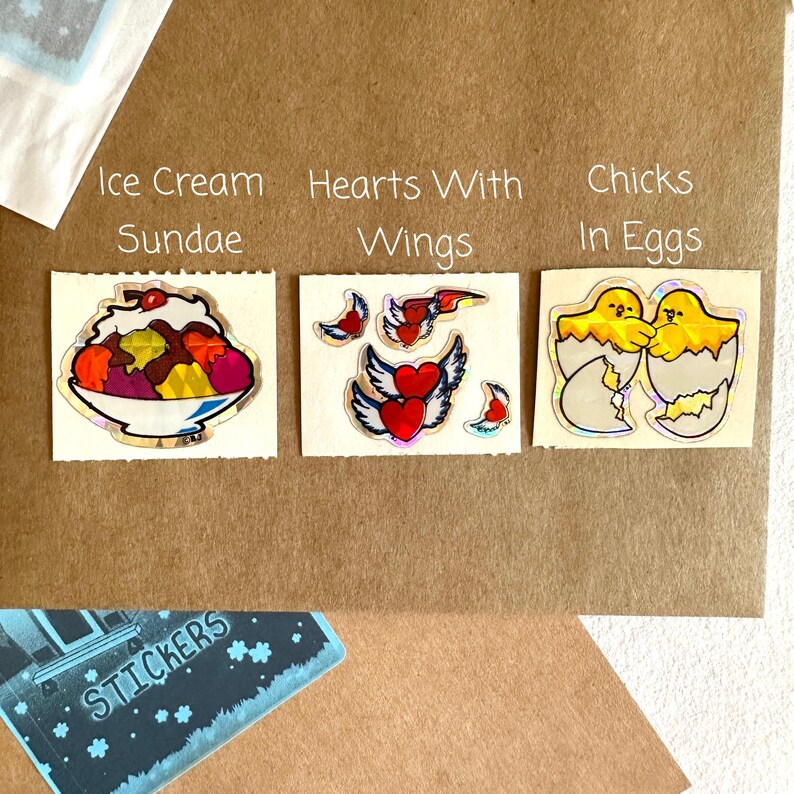 May include: Three sticker sheets with holographic designs. The first sheet features an ice cream sundae, the second sheet features hearts with wings, and the third sheet features chicks in eggs.