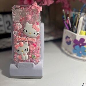 Op de afbeelding: Een doorzichtige telefoonhoes versierd met roze Hello Kitty-figuren, strikken en snoepjes. De hoes is versierd met roze kralen en een roze strik aan de bovenkant. De woorden "Hello Kitty" zijn zichtbaar op de hoes.