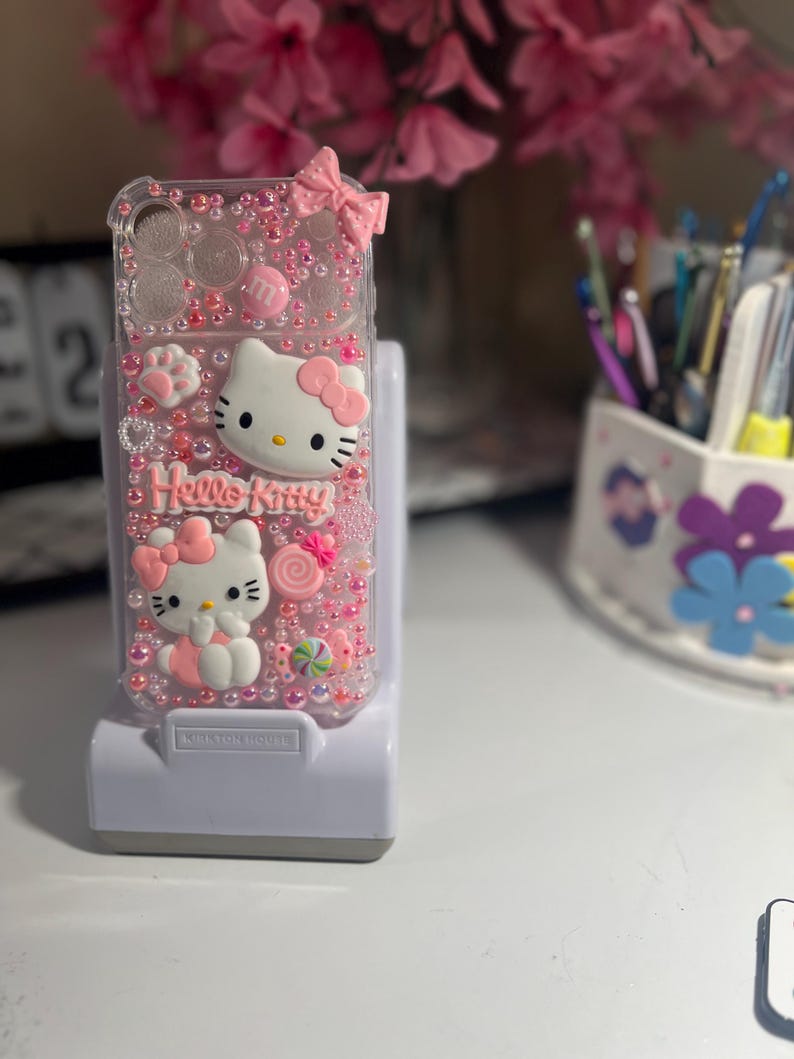 Op de afbeelding: Een doorzichtige telefoonhoes versierd met roze Hello Kitty-figuren, strikken en snoepjes. De hoes is versierd met roze kralen en een roze strik aan de bovenkant. De woorden "Hello Kitty" zijn zichtbaar op de hoes.