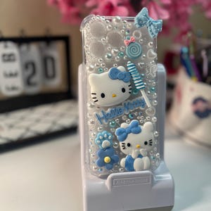 Op de afbeelding: Transparante telefoonhoes met Hello Kitty-figuren, blauwe strikken en een lolly. De hoes is versierd met imitatieparels en de woorden "Hello Kitty". De hoes staat op een witte standaard.