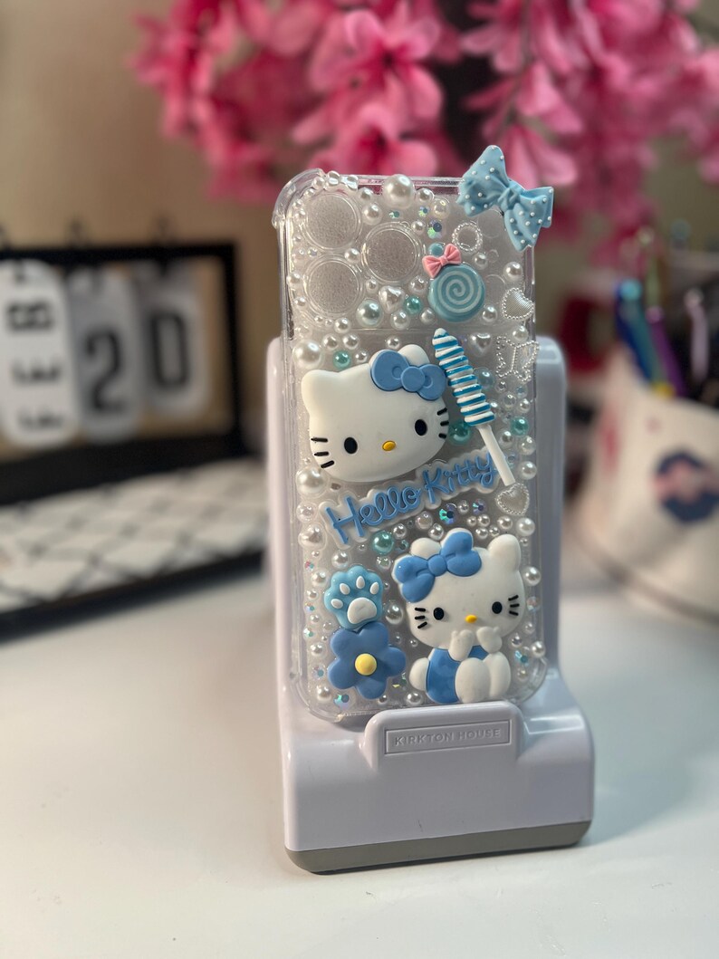 Op de afbeelding: Transparante telefoonhoes met Hello Kitty-figuren, blauwe strikken en een lolly. De hoes is versierd met imitatieparels en de woorden "Hello Kitty". De hoes staat op een witte standaard.