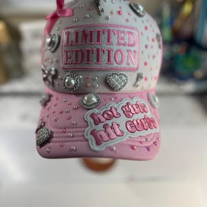 May include: Pink and white baseball cap with the text "LIMITED EDITION" embroidered in pink. The cap is embellished with pearls, rhinestones, and small charms. The brim features the text "hot girls hit curbs".