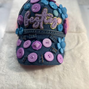 May include: A dark blue denim baseball cap adorned with various purple, light blue, and pink buttons, flower-shaped embellishments, and a central fuzzy purple patch featuring the word besties in cursive script with a gold glitter border.