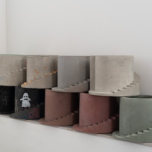 Peut inclure: Une rangée de neuf pots en béton avec un design d'escalier en spirale. Les pots sont de différentes couleurs, notamment noir, gris, vert et marron.