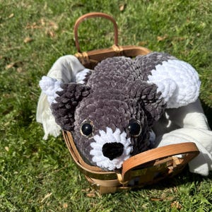 May include: A gray and white crocheted stuffed animal in the shape of a dog, lying in a wicker basket. The dog has large black eyes and a black nose.