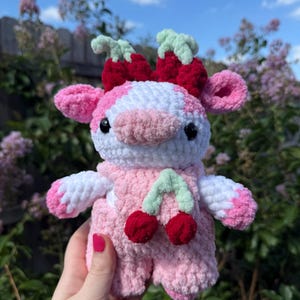May include: A plush cow toy made of white and pink yarn with black safety eyes. The cow features a pair of red cherries with light green stems on its head and a pair of red cherries with light green stems attached to its pink crocheted overalls.