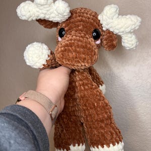 May include: A plush crochet moose toy made from soft brown yarn with white textured antlers and white accents on the hooves. The stuffed animal features large black safety eyes and small pink embroidered details beneath the eyes on its face.