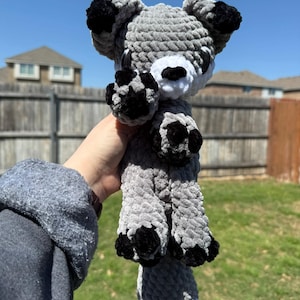 May include: A gray and black crocheted fox plush toy with white accents. The toy has a long tail and black paws.
