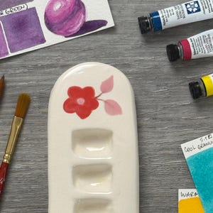 Puede incluir: Una paleta de acuarela de cerámica blanca con un diseño de flor roja y cinco pozos. Tubos de acuarela, muestras de pintura y pinceles están dispuestos alrededor de la paleta sobre una superficie de madera. Las muestras de pintura tienen el texto "HANSA YELLOW LIGHT COOL D.S."