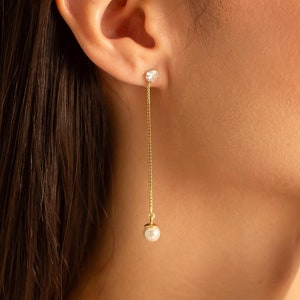 Op de afbeelding: Een paar gouden oorhangers met een klein, helder steentje bovenaan en een witte parel onderaan.