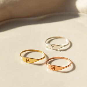 Può includere: Tre anelli in oro, argento e oro rosa con una fascia piatta di forma ovale. Ogni anello ha una singola lettera incisa sulla fascia: "B", "Y" e "M".