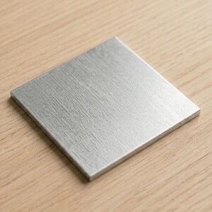 Puede incluir: Un posavasos cuadrado de metal cepillado con una superficie texturizada. El posavasos es plateado y se encuentra sobre una superficie de madera marrón claro. Diseño sencillo y moderno.