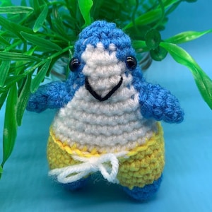 May include: A crocheted penguin wearing yellow swim trunks. The penguin has blue and white fur and black eyes.