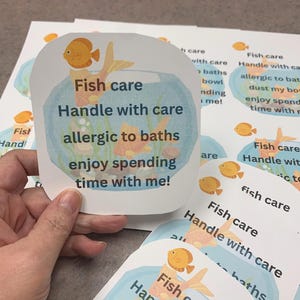 Puede incluir: Una mano sostiene una tarjeta impresa con un pez dorado de dibujos animados en un cuenco. La tarjeta dice "Fish care, Handle with care, allergic to baths, enjoy spending time with me!" Se ven otras tarjetas.