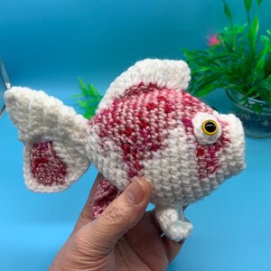 May include: A crocheted goldfish toy with pink and white yarn. The toy has a white body with pink spots and a yellow eye.