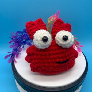 May include: A handmade crochet crab in bright red, featuring large white eyes with black pupils and a smiling mouth. The crab is adorned with decorative purple and pink floral accents, set against a vibrant blue backdrop.