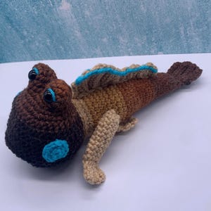 May include: A crocheted mudskipper toy featuring a dark brown head, a tan midsection, and a dark brown tail. The creature has two black plastic eyes with bright blue accents, a blue-trimmed dorsal fin, and two light tan pectoral fins.