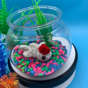 May include: A white crocheted fish with a red fin and black eyes sits in a glass bowl filled with colorful pebbles. The bowl is on a black and white base.