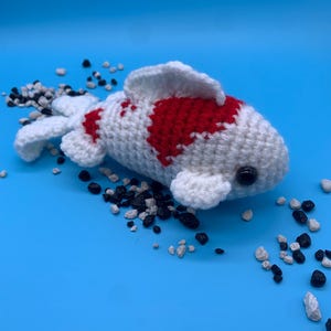 May include: A handmade crochet koi fish in white with red markings and black eyes. The fish is surrounded by small black and white pebbles on a blue background. The fish has fins and a tail.